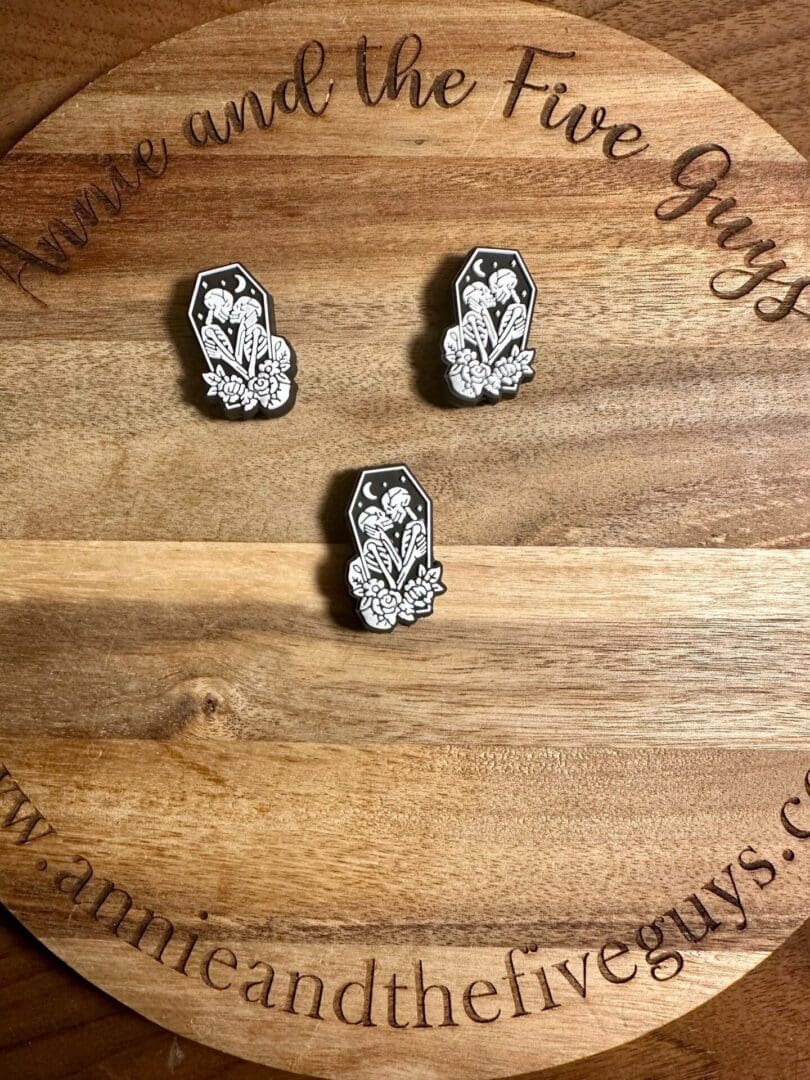 Three Kissing skeletons in a coffin silicone focal beads featuring white floral designs on a wood grain background with the text "Annie and the Five Guys.