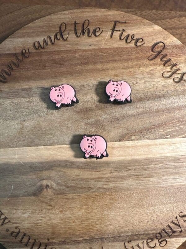 Three pink pig stickers laid out on a wooden surface with cursive text partially visible around the edge.