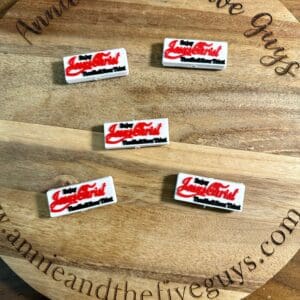 Five rectangular pins with red and black text reading "Lady Luscious Lips" displayed on a circular wooden board with the text "Annie and the Five Guys" around the edge.