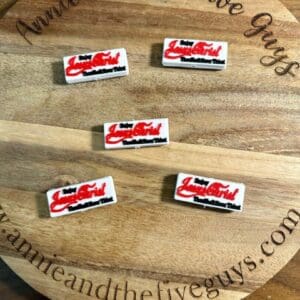 Five rectangular pins with red and black text reading "Lady Luscious Lips" displayed on a circular wooden board with the text "Annie and the Five Guys" around the edge.