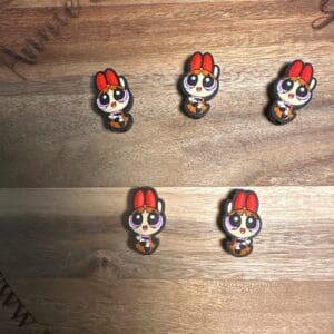 Five small, cartoonish pins featuring a character with red hair and big eyes displayed on a wooden surface with text partially visible around the edge.