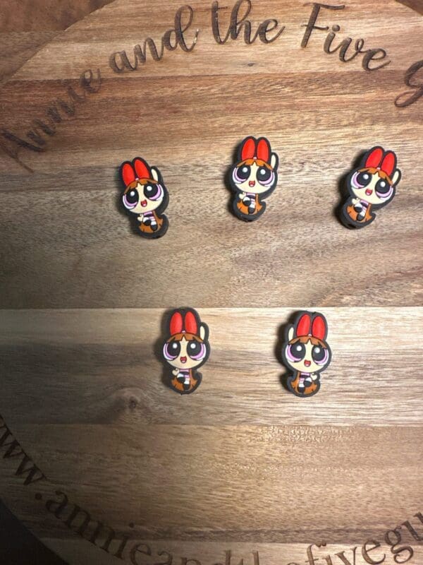 Five small, cartoonish pins featuring a character with red hair and big eyes displayed on a wooden surface with text partially visible around the edge.