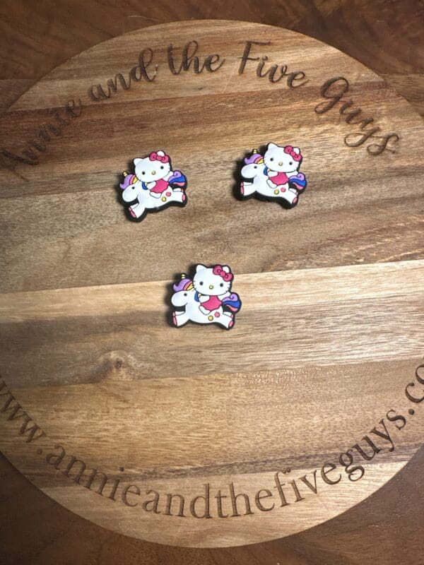 Three White HK riding a unicorn pins with pink bows, wings, and rainbow tails are positioned on a round wooden coaster featuring the text "Annie and the Five Guys" and a website URL.