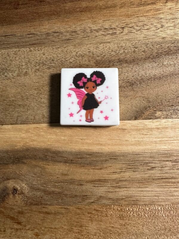 Tile featuring a cartoon fairy with dark skin, curly hair styled in puffs with pink bows, a black dress, pink wings, and holding a magic wand, set against a wooden background is replaced with African American girl, women, Juneteenth silicone focal bead.