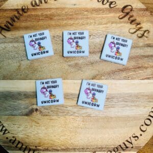 Five square tiles on a wooden surface, each featuring an illustration of a unicorn with the text "I'm not your ordinary unicorn." The surface has the text "Annie and the Five Guys" engraved around the edge.