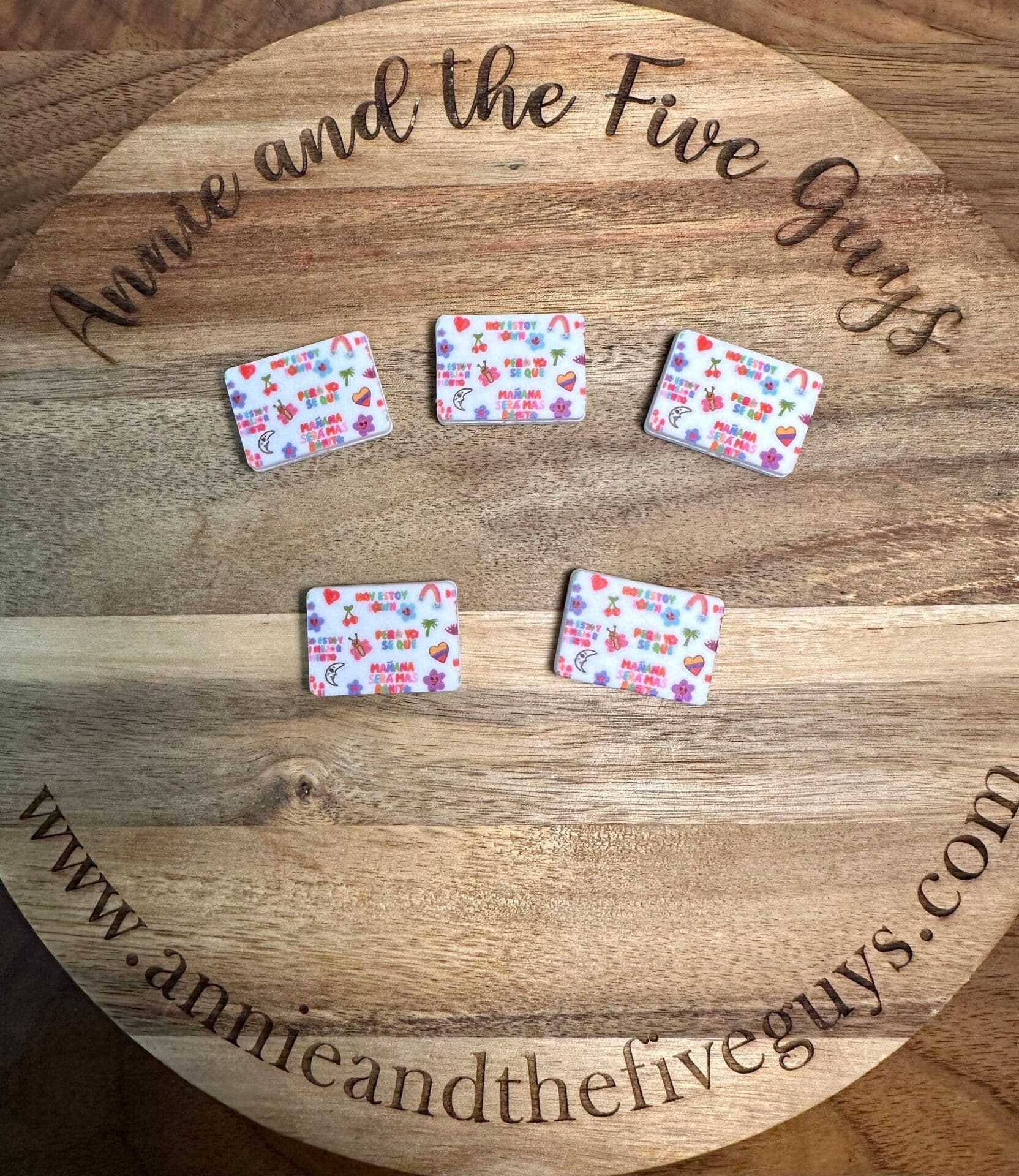 Five small decorated rectangular items on a wooden surface with text "Annie and the Five Guys" and a website link around the edges.