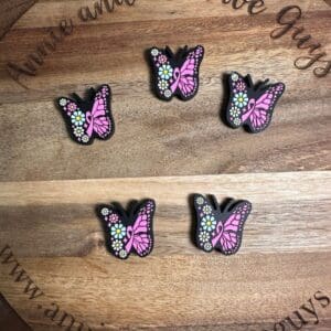 Five pink and black butterfly clips with colorful flower details are displayed on a wooden surface. The surface has the text "Annie and the Five Guys" and the website "www.annieandthefiveguys.com.