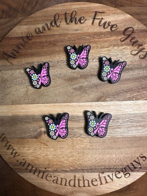 Five pink and black butterfly clips with colorful flower details are displayed on a wooden surface. The surface has the text "Annie and the Five Guys" and the website "www.annieandthefiveguys.com.