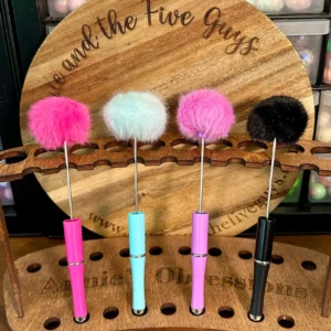 Four colorful fluffy beadable pens with pom pom (pink, blue, light pink, and black) are displayed vertically in a wooden holder, with "Five and the Five Guys Annie Obsessions" engraved on the back.