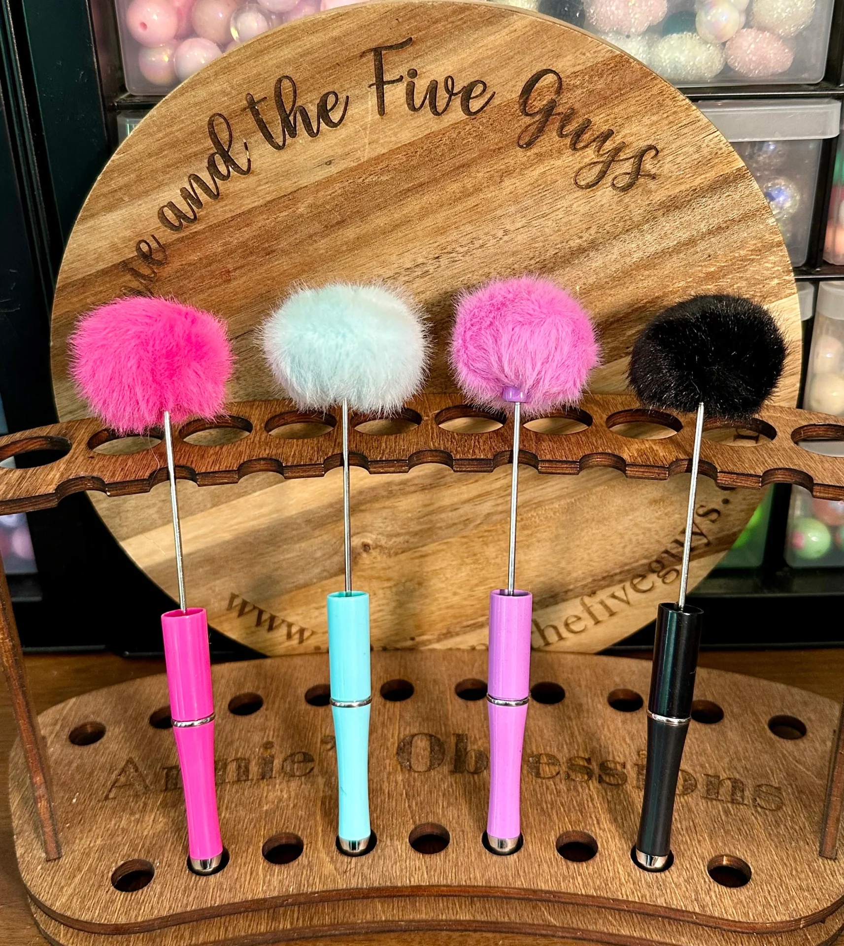 Four colorful fluffy beadable pens with pom pom (pink, blue, light pink, and black) are displayed vertically in a wooden holder, with "Five and the Five Guys Annie Obsessions" engraved on the back.