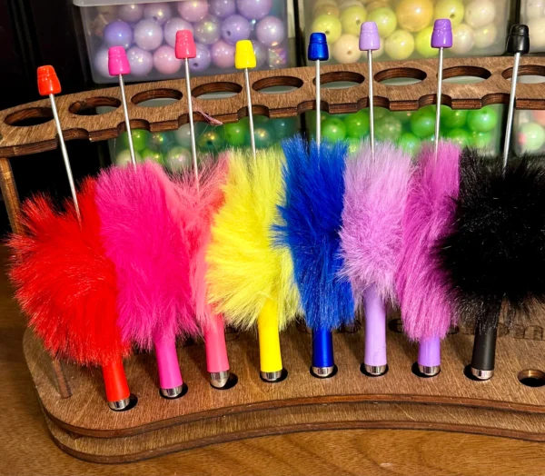 Brightly colored Pen, fluffy, plastic, Beadable in red, pink, yellow, blue, purple, and black are neatly arranged in a wooden stand, with plastic balls in containers in the background.