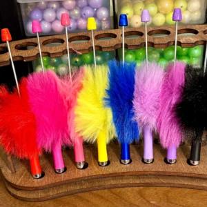 Brightly colored Pen, fluffy, plastic, Beadable in red, pink, yellow, blue, purple, and black are neatly arranged in a wooden stand, with plastic balls in containers in the background.