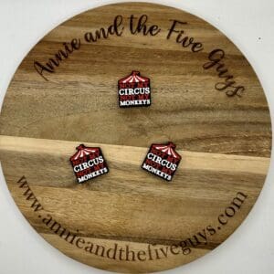 A round wooden board engraved with "Annie and the Five Guys" and "www.annieandthefiveguys.com" displaying three square pins that read "Not my circus, not my monkeys" with a red circus tent graphic.