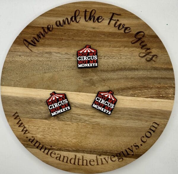 A round wooden board engraved with "Annie and the Five Guys" and "www.annieandthefiveguys.com" displaying three square pins that read "Not my circus, not my monkeys" with a red circus tent graphic.