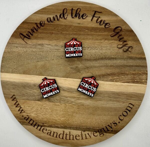 A round wooden board engraved with "Annie and the Five Guys" and "www.annieandthefiveguys.com" displaying three square pins that read "Not my circus, not my monkeys" with a red circus tent graphic.