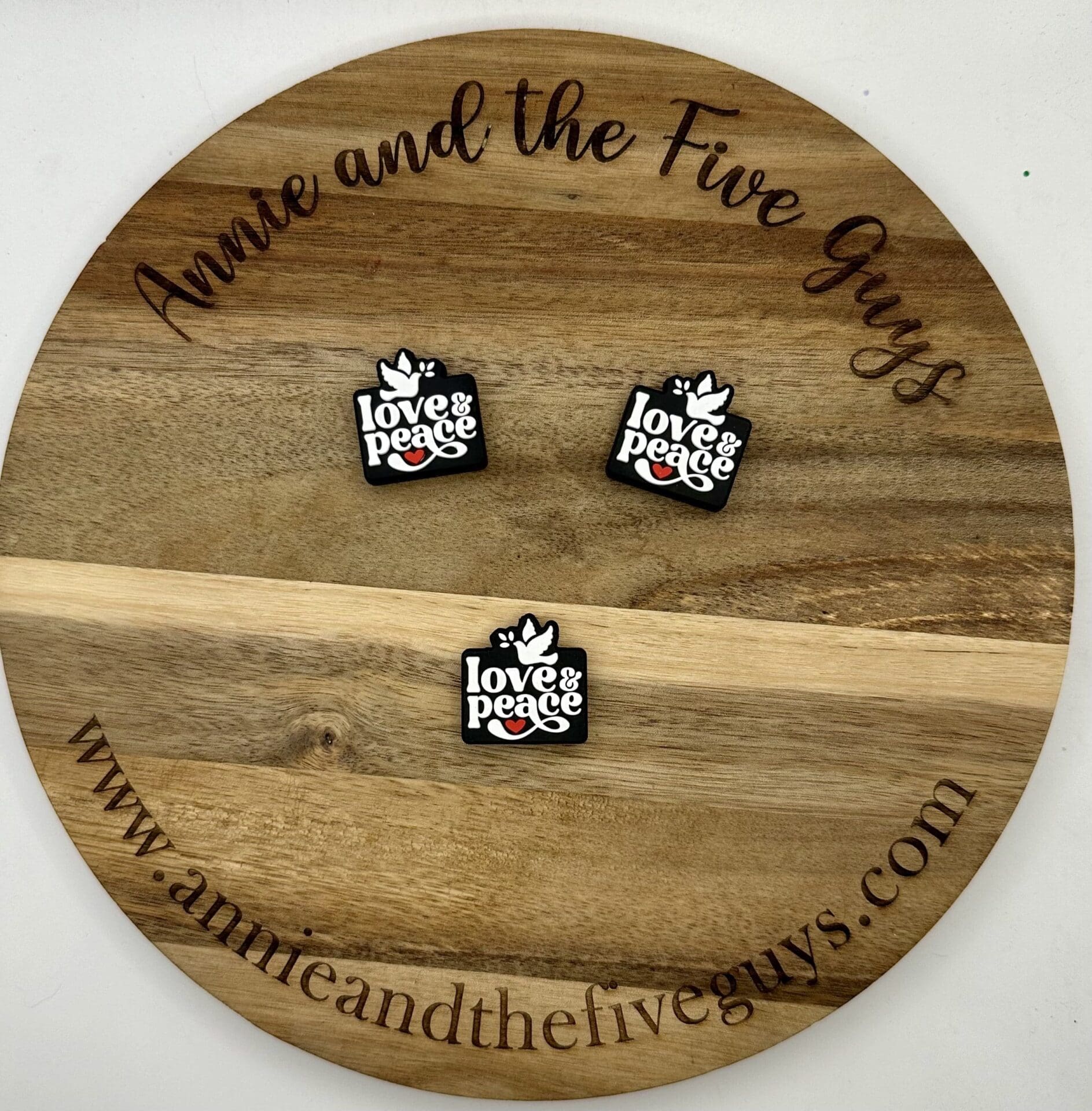 Wooden circular board engraved with "Annie and the Five Guys" and website link, displaying three square pins with the text "love & peace" and a dove illustration.