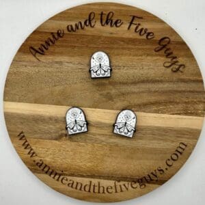 A wooden circular board with the text "Annie and the Five Guys" engraved along its edge. Three small, intricately designed pieces are placed on the board. The website annieandthefiveguys.com is also visible.