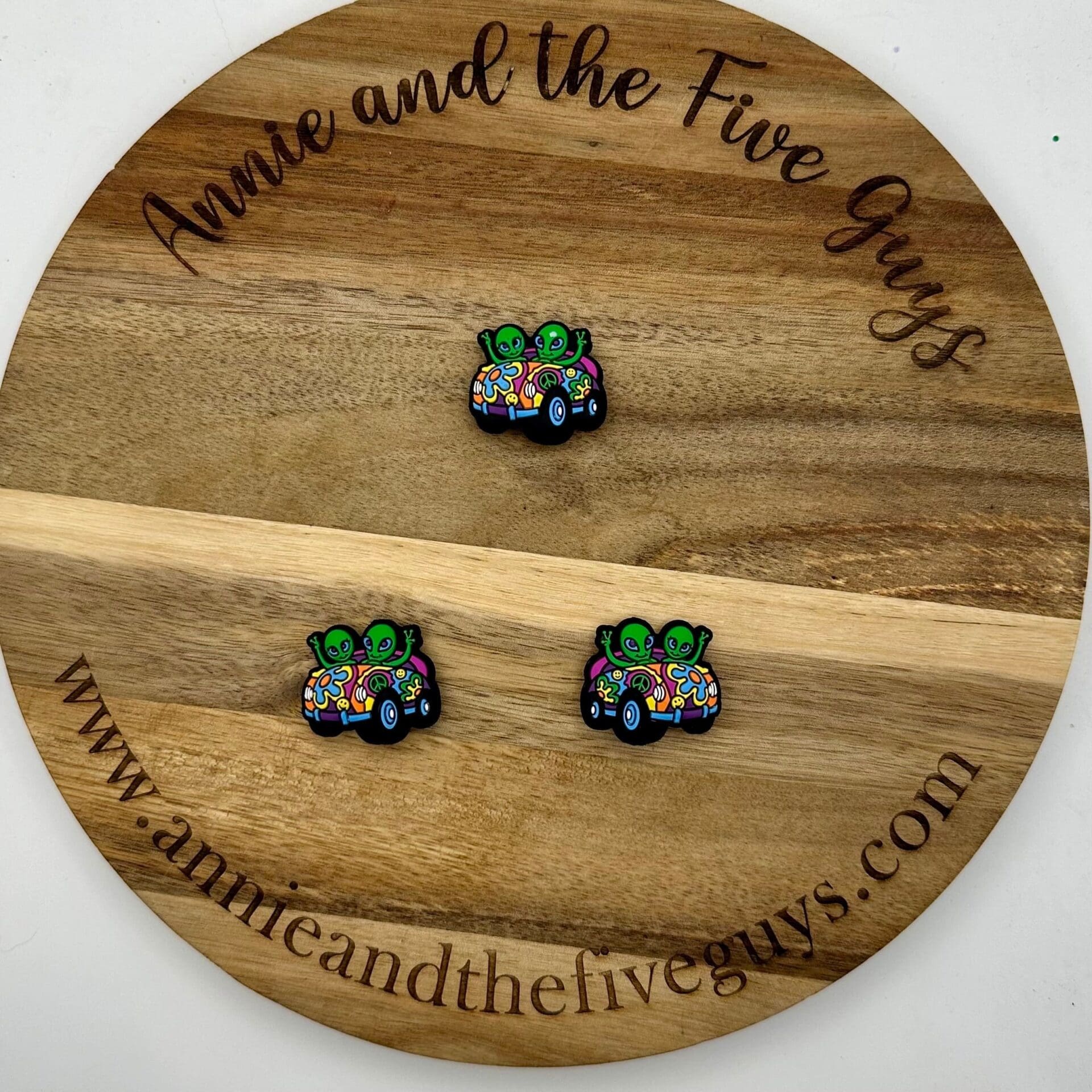 A round wooden board with "Annie and the Five Guys" engraved, featuring three pins of cartoon aliens driving colorful cars.