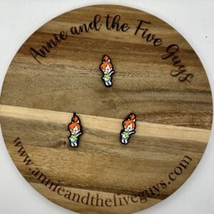 Three small enamel pins featuring a character with orange hair, placed on a round wooden board with the inscription "Annie and The Five Guys" and the website "www.annieandthefiveguys.com.