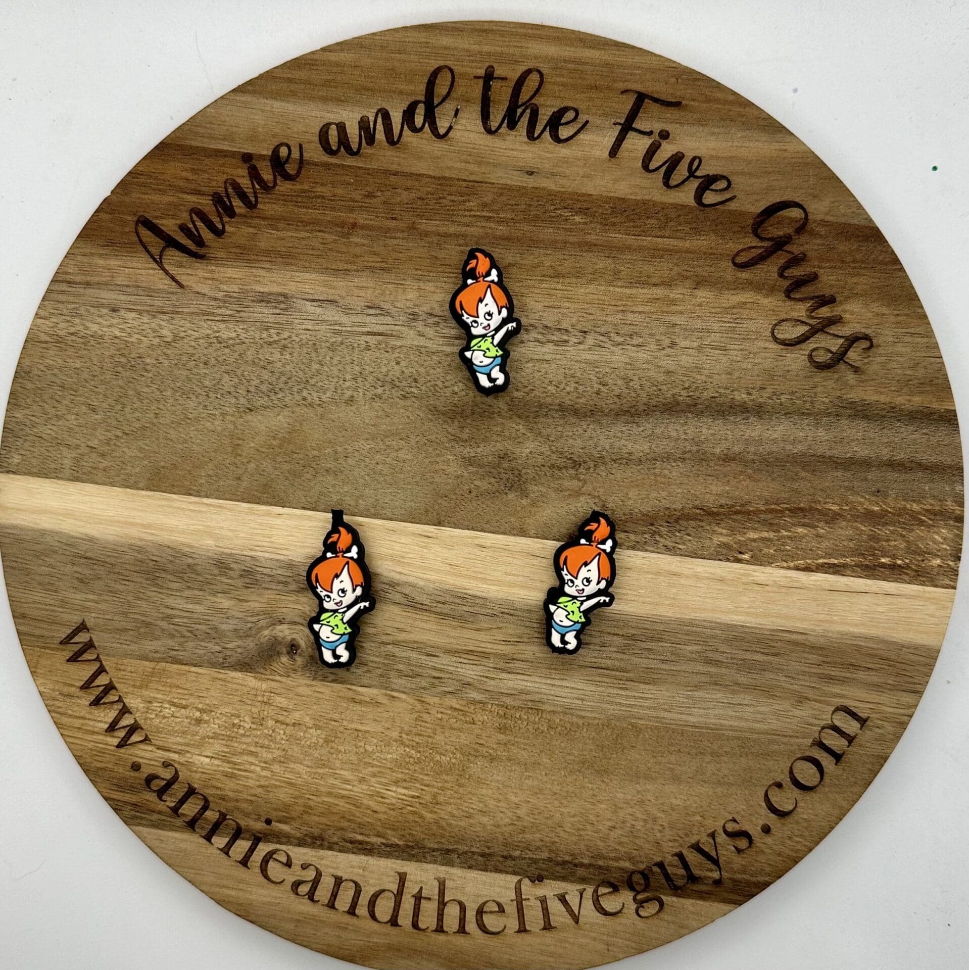 Three small enamel pins featuring a character with orange hair, placed on a round wooden board with the inscription "Annie and The Five Guys" and the website "www.annieandthefiveguys.com.