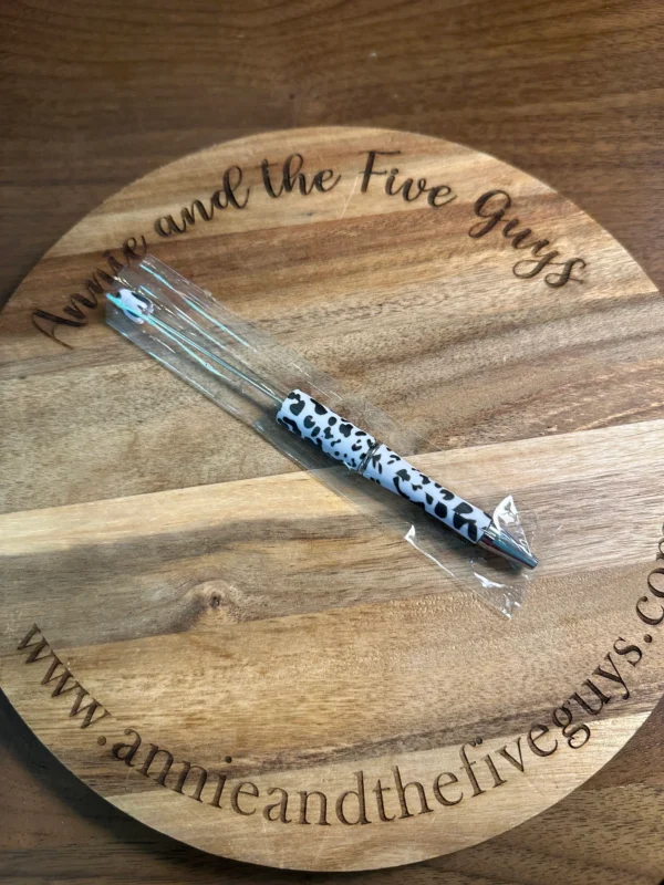 A Printed Beadable pen rests on a circular wooden board engraved with "Annie and the Five Guys" and the corresponding website URL.