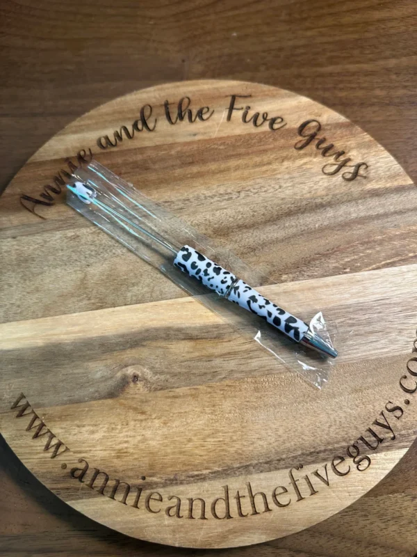 A Printed Beadable pen rests on a circular wooden board engraved with "Annie and the Five Guys" and the corresponding website URL.