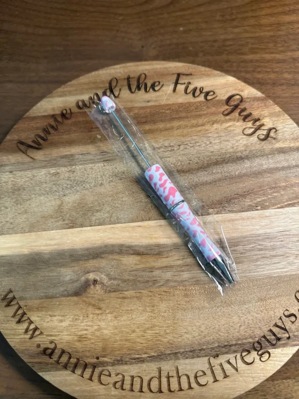 A Printed Beadable pen wrapped in plastic lies on a wooden round board with text engraved around the edge.