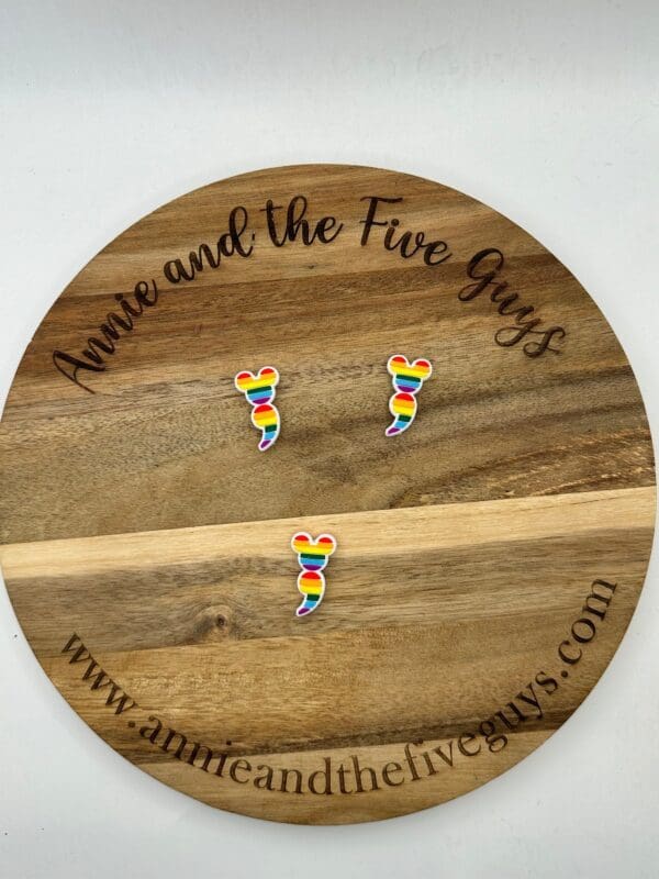 Three rainbow-colored mouse ear-shaped pins are placed on a round wooden board with the text "Annie and the Five Guys" and the website "www.annieandthefiveguys.com.