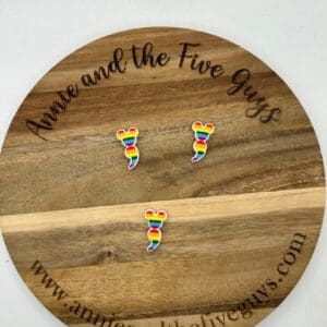 Three rainbow-colored mouse ear-shaped pins are placed on a round wooden board with the text "Annie and the Five Guys" and the website "www.annieandthefiveguys.com.