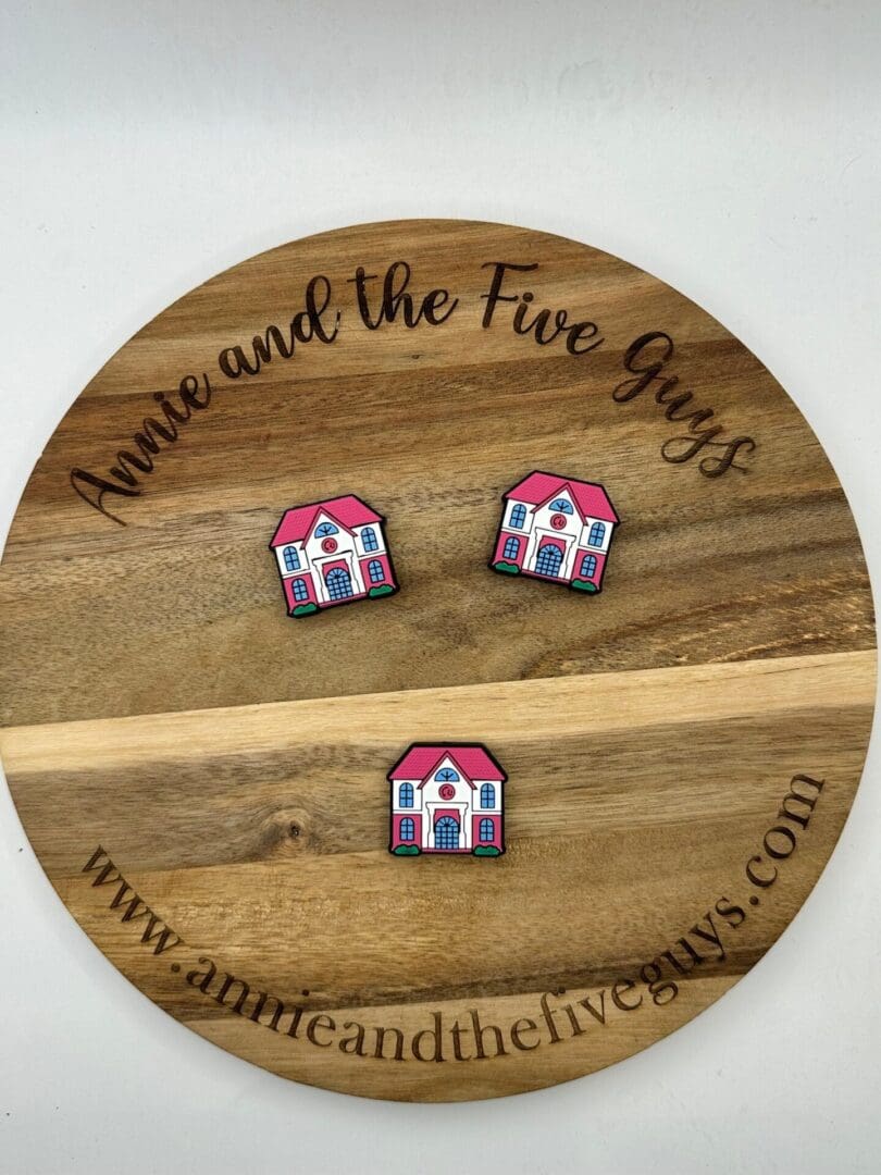 A round wooden board with the text "Annie and the Five Guys" displays two pairs of small, house-shaped pins. The website URL www.annieandthefiveguys.com is also visible on the board.