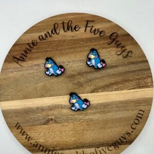 Three blue and pink enamel pins featuring a character with a sad expression displayed on a wooden board with the text "Annie and the Five Guys" and the website www.annieandthefiveguys.com.