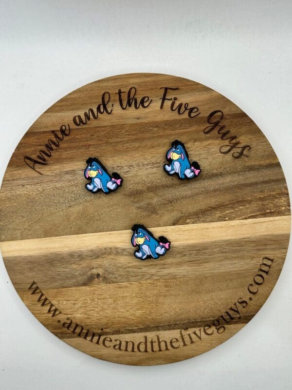 Three blue and pink enamel pins featuring a character with a sad expression displayed on a wooden board with the text "Annie and the Five Guys" and the website www.annieandthefiveguys.com.