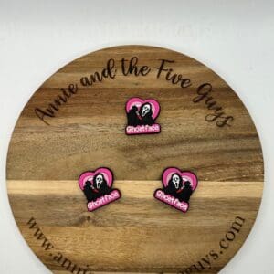 A round wooden board with the text "Annie and the Five Guys" engraved on the rim. Three pink and black "Ghostface" pins are placed on the board. The website "www.annieandthefiveguys.com" is at the bottom.