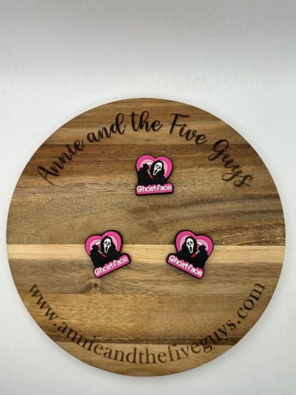 A round wooden board with the text "Annie and the Five Guys" engraved on the rim. Three pink and black "Ghostface" pins are placed on the board. The website "www.annieandthefiveguys.com" is at the bottom.