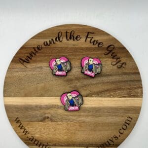 Three Jason silicone focal beads with a graphic of a character holding a sack and a heart are displayed on a round wooden board with the text "Annie and the Five Guys" engraved on it.