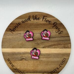 A wooden circular board with the text "Annie and the Five Guys" displays three identical pins with a figure and the name "Freddie." The website address is visible at the bottom.