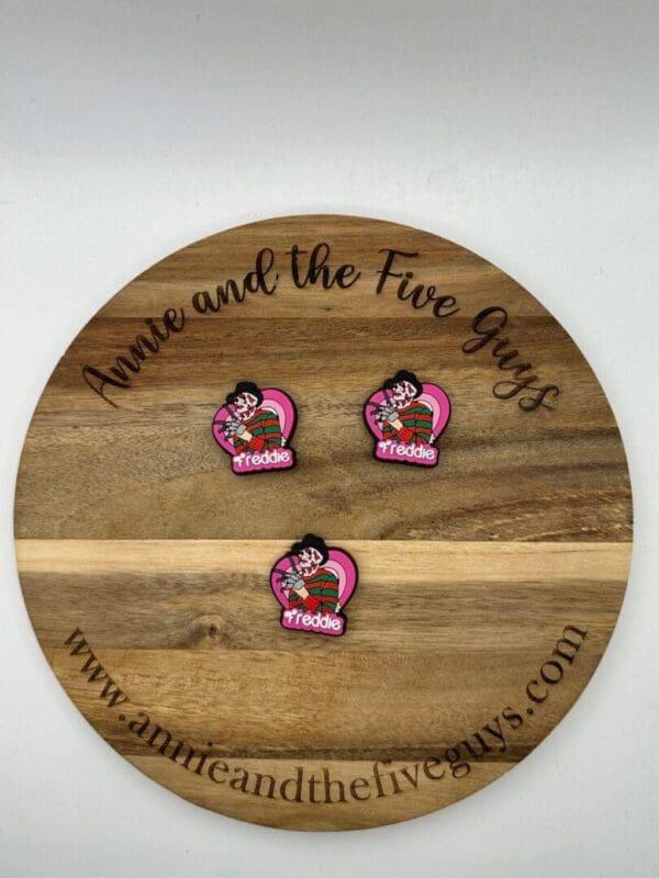 A wooden circular board with the text "Annie and the Five Guys" displays three identical pins with a figure and the name "Freddie." The website address is visible at the bottom.