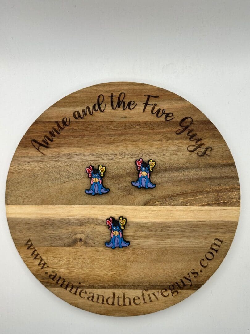 A round wooden board with the text "Annie and the Five Guys" displays three colorful pins shaped like blue slugs with butterfly wings on their heads. Website URL is visible at the bottom of the board.