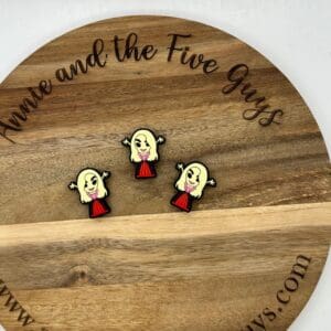 Wooden circular board with three enamel pins of a blonde character in a red dress. Text on the board reads “annie and the Five Guys.”.