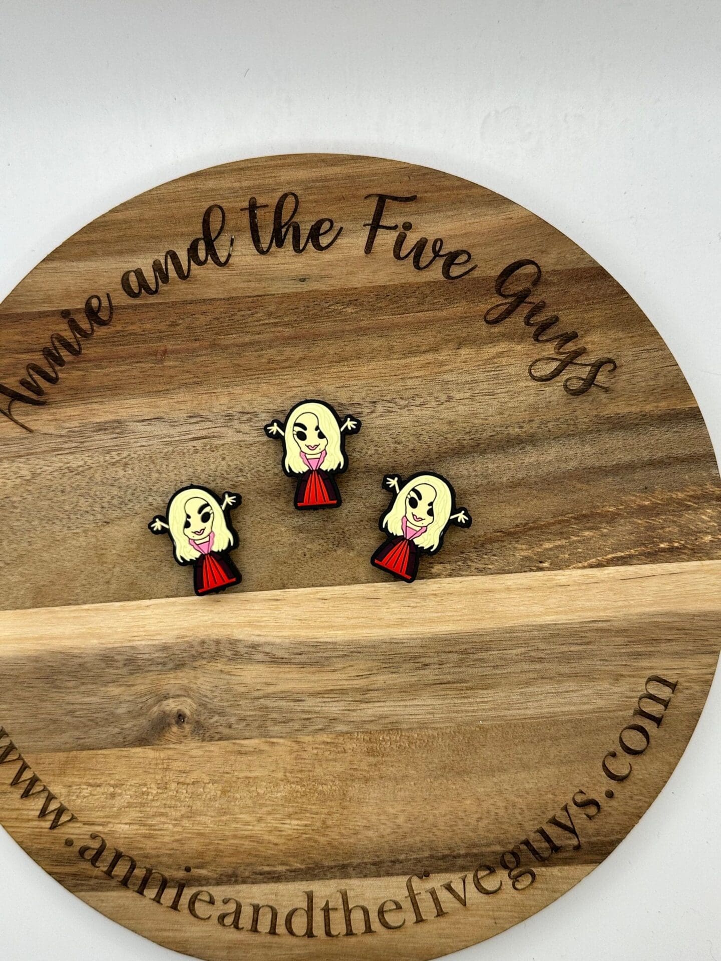 Wooden circular board with three enamel pins of a blonde character in a red dress. Text on the board reads “annie and the Five Guys.”.