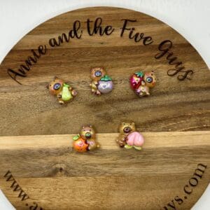 A wooden disc with "Annie and the Five Guys" written on it, displaying five decorated, small Fruit bear acrylic focal beads. Each bead is holding a different fruit or dessert.