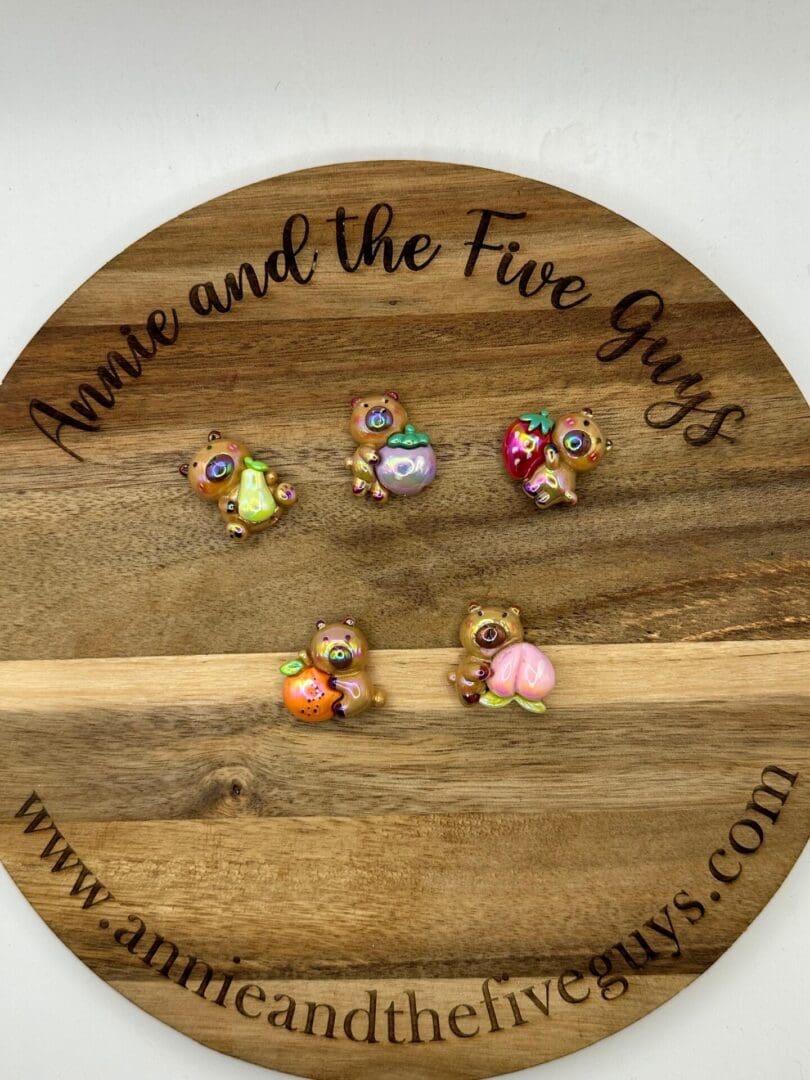 A wooden disc with "Annie and the Five Guys" written on it, displaying five decorated, small Fruit bear acrylic focal beads. Each bead is holding a different fruit or dessert.