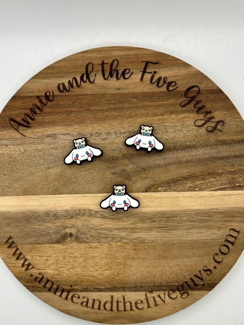 A circular wooden board with three small pins on it, each depicting a panda with wings. The board is engraved with the text "Cinnamon bear silicone focal bead" and the website "www.annieandthefiveguys.com".