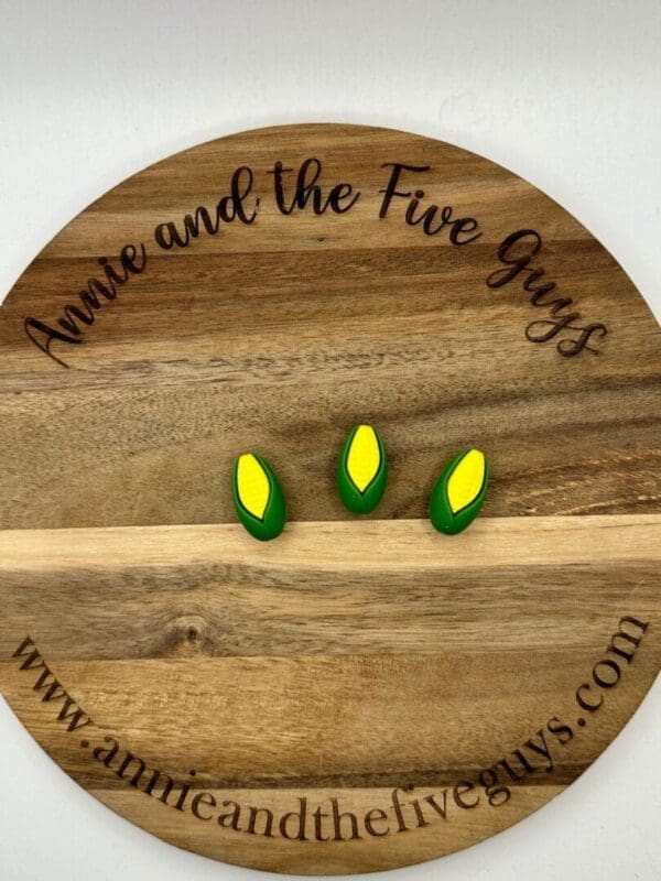 A round wooden board with three small ear of corn magnets. The board is labeled "Annie and the Five Guys" with the website "www.annieandthefiveguys.com".