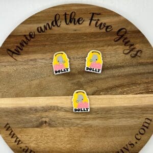 Wooden round board with black cursive text displaying the website “Annie and the Five Guys.” Three cartoon stickers of a blonde woman with the name “Dolly” are placed on the board.