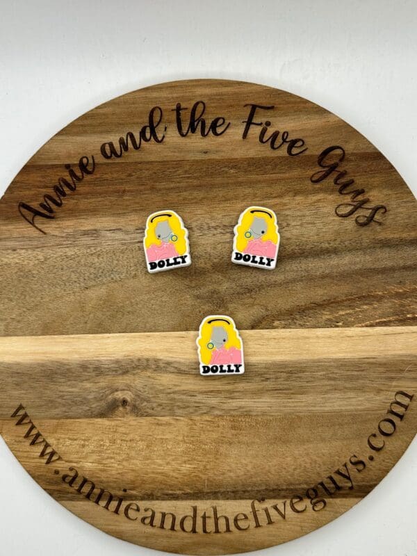 Wooden round board with black cursive text displaying the website “Annie and the Five Guys.” Three cartoon stickers of a blonde woman with the name “Dolly” are placed on the board.