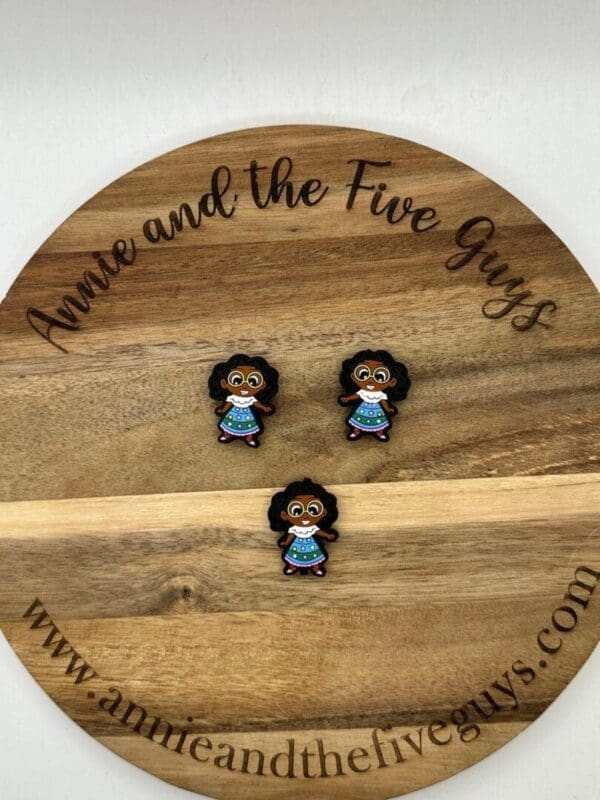 Three small figurines of a child with curly hair and a blue dress are displayed on a round wooden plaque that reads "Annie and the Five Guys" around the edge.