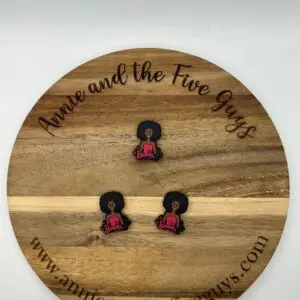 A round wooden board with the text "Annie and the Five Guys" holds three small pins of a figure with an afro hairstyle, and a pink top with "Barbie" written on it. Website URL is displayed at the bottom.