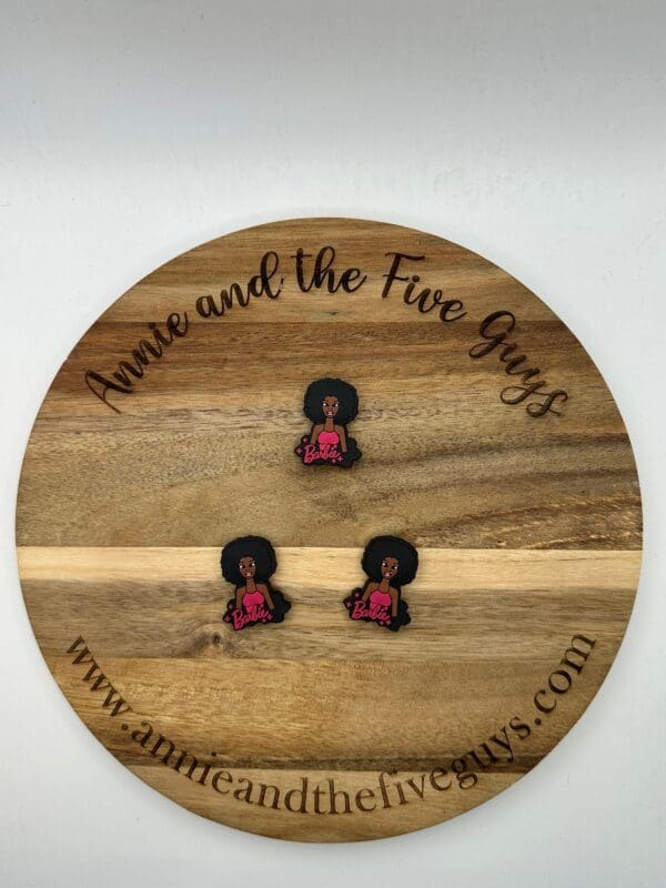 A round wooden board with the text "Annie and the Five Guys" holds three small pins of a figure with an afro hairstyle, and a pink top with "Barbie" written on it. Website URL is displayed at the bottom.