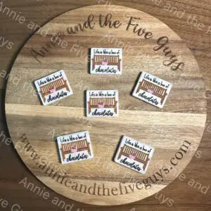 A round wooden board with six stickers, each bearing the text "Exclusive ‘Life is like a box of chocolates’ silicone focal bead" and an illustration of a bench. The text "Annie and the Five Guys" is on the board's edge and background.